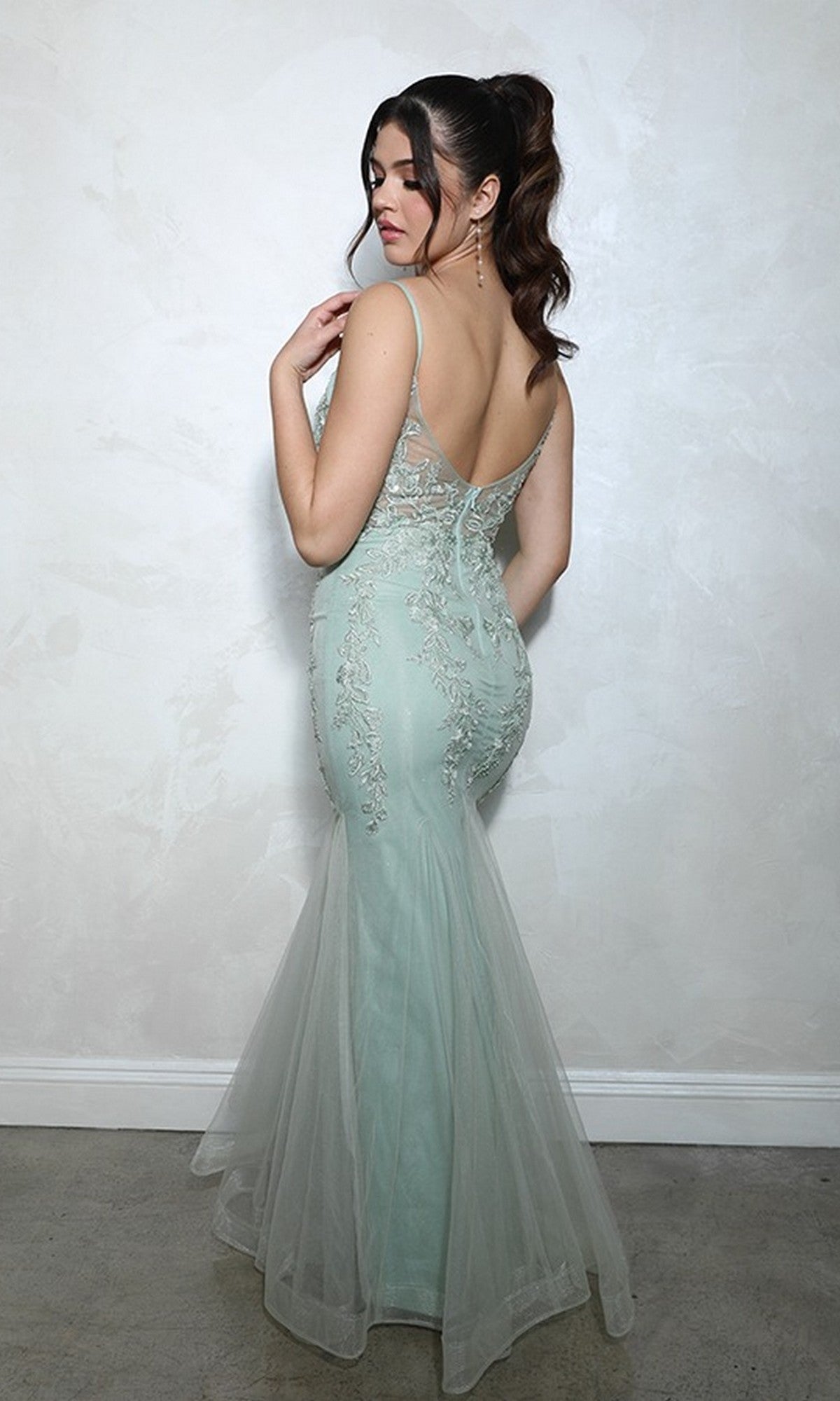 A woman models the Long Prom Dress: Eureka 9998, a light blue gown with embroidered beaded floral details, standing against a plain textured wall while looking over her shoulder.